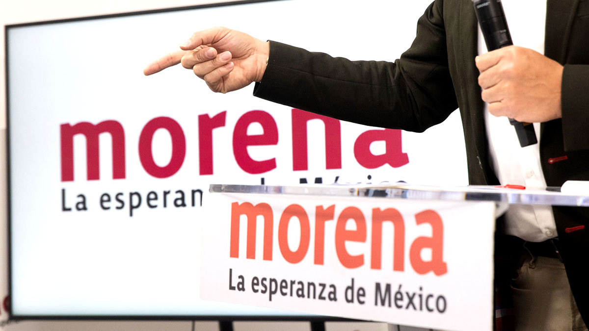 Elecciones 2024 Morena: Hombre en atril con logo de Morena, igual en una pantalla tras él.